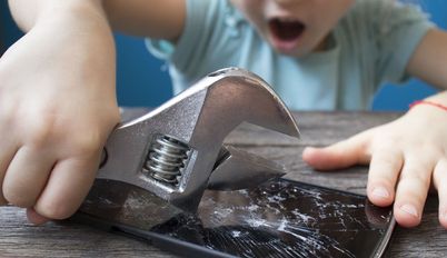 Ein Kind schlägt schwungvoll mit einem Werkzeug auf ein Mobiltelefon, das auf dem Tisch liegt