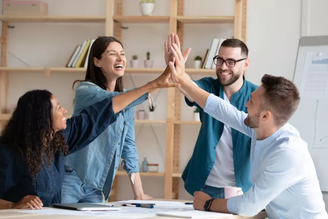 4 junge Leute in einem modernen Büro, die am Tisch sitzen und sich lachend High-Five geben