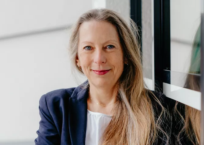 Beraterin Simone Gehr im blauen Anzug lehnt an einem Glasfenster mit Spiegelung