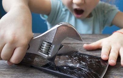 Ein Kind schlägt schwungvoll mit einem Werkzeug auf ein Mobiltelefon, das auf dem Tisch liegt