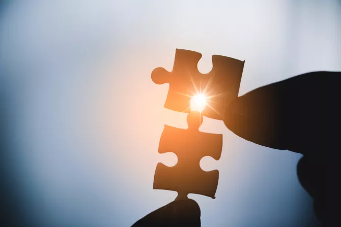 Zwei Finger halten zwei Puzzlestücke gegeneinander. Durch eine Lücke strahlt die gelbe Sonne